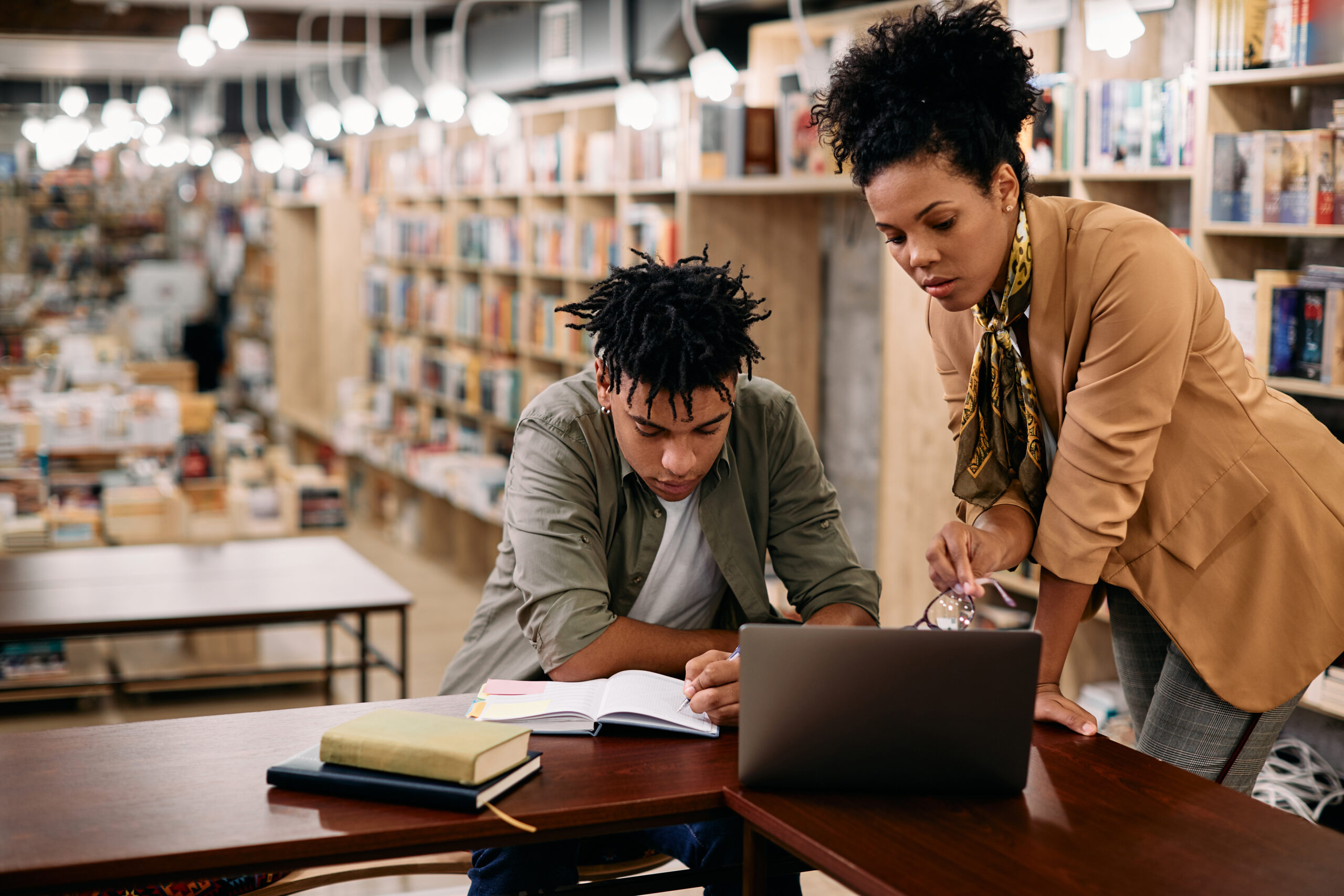 A mentor guiding a student over an open notebook and laptop in a library, symbolizing the intellectual partnership of The Ars Cogitans™ and Vocation.