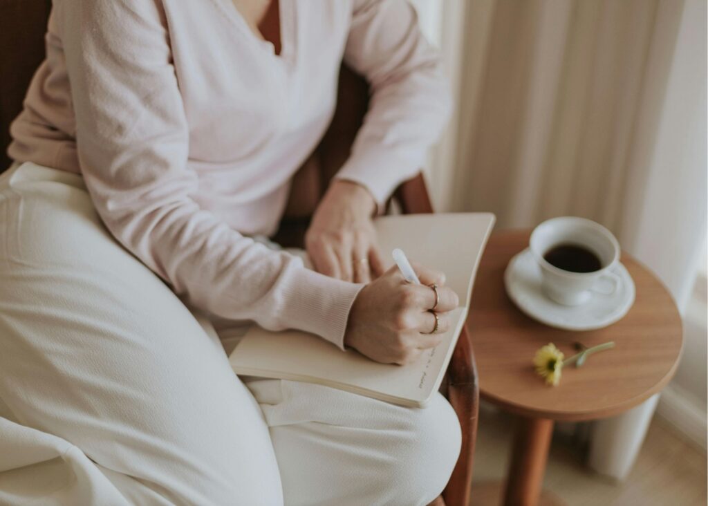 Woman writing in a journal beside a cup of coffee on a small table in soft morning light.