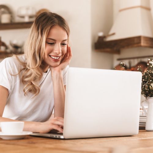 A woman smiling while working on her laptop in a cozy kitchen, symbolizing peaceful winter homeschool planning.